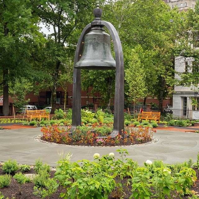 Large bell suspended by an arch surrounded by footpaths and flowerbeds. 