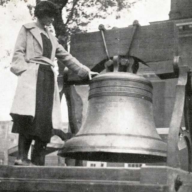Black and white photo of a young woman standing on a platform next to the Women's Liberty Bell.