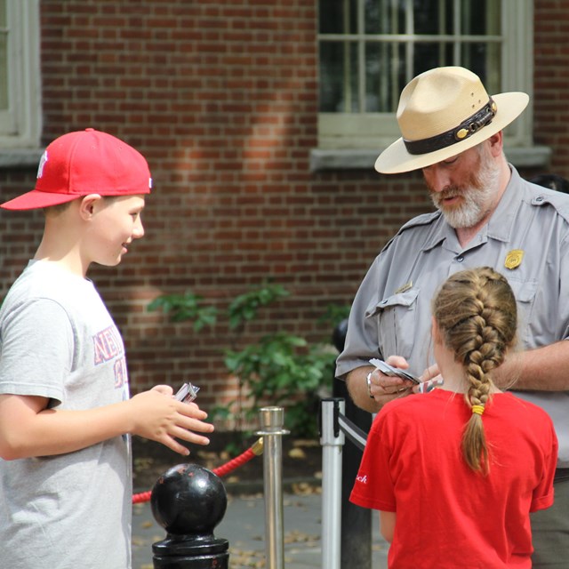 Color photo of a male park ranger standing outdoors, handing trading cards to a boy and girl.
