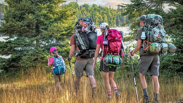 A family with three adults and one child hikes in a forested meadow with large backpacks.