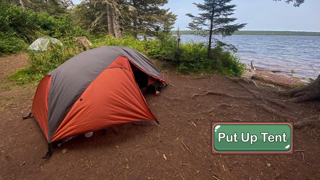 A red and gray tent pitched at a lakeside campsite.