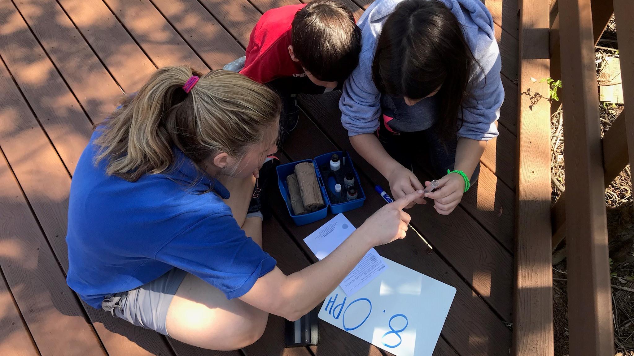 A young woman points to a test tube. Two kids look at the test tube. Small white board on ground.