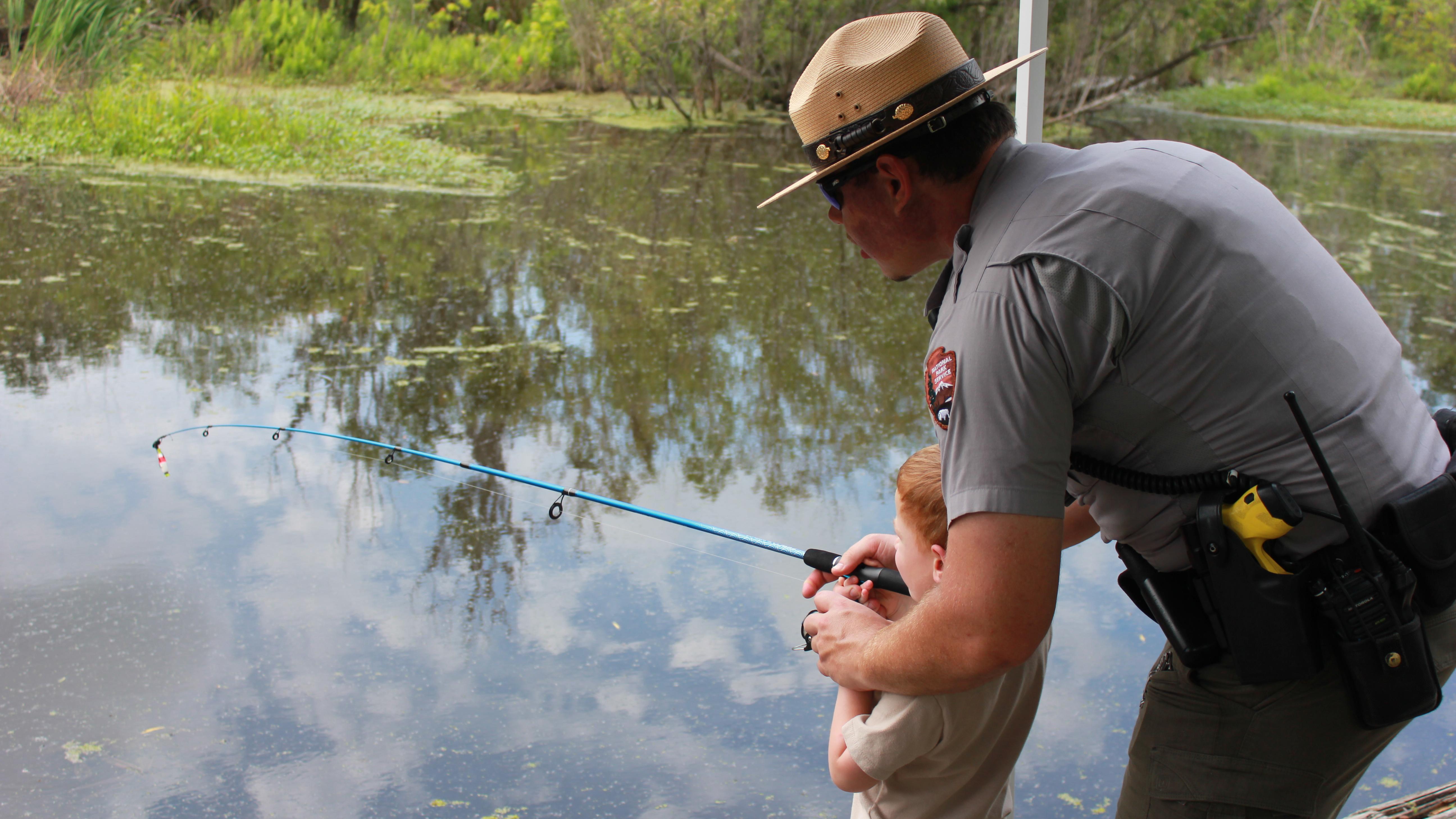 A law enforcement ranger helps a kid with a fishing pole next to a bayou.