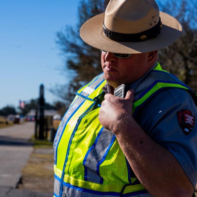 A law enforcement ranger in a flat hat & wearing a safety vest talks into a radio