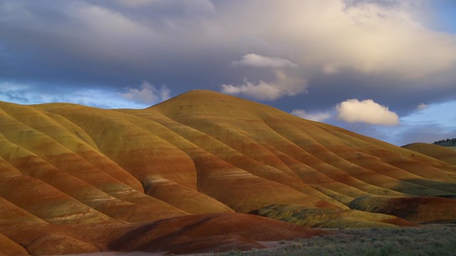 A line of tan and red stripped hills lit up by the sun below a cloudy sky.