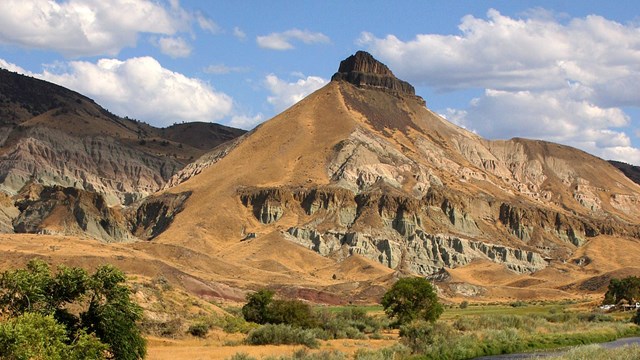 A pyramid-shaped hill with a craggy, black top reaches into the sky with grasslands and river below.