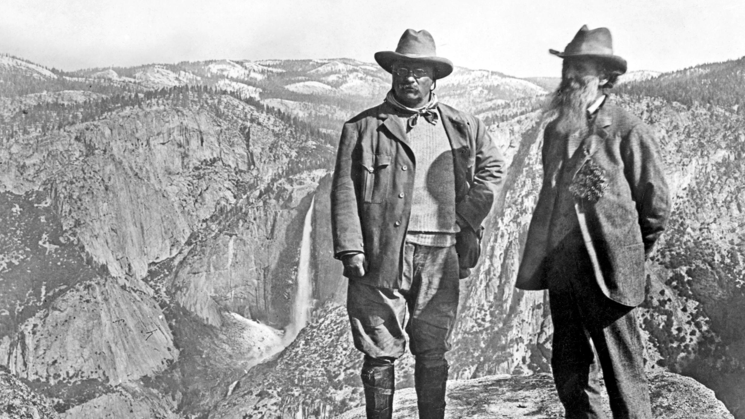 Two men in suits stand on a mountain. One beard, one mustache and glasses. 