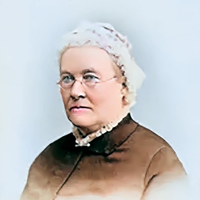 A senior woman with short hair, glasses and wearing a cap and dress is posing for a photo.