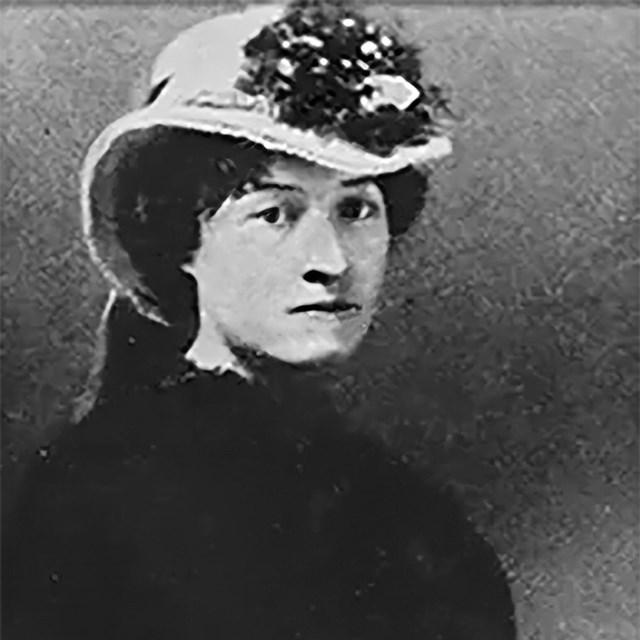 A middle-aged woman in a hat and dress poses for the camera. 