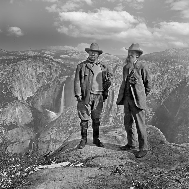 Two older men stand on a rock with mountains behind them. 