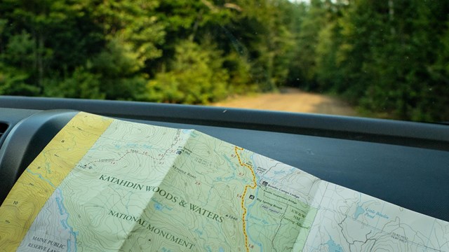A green map sits on a dashboard. Some map lines and words are visible. A dirt road extends outside.