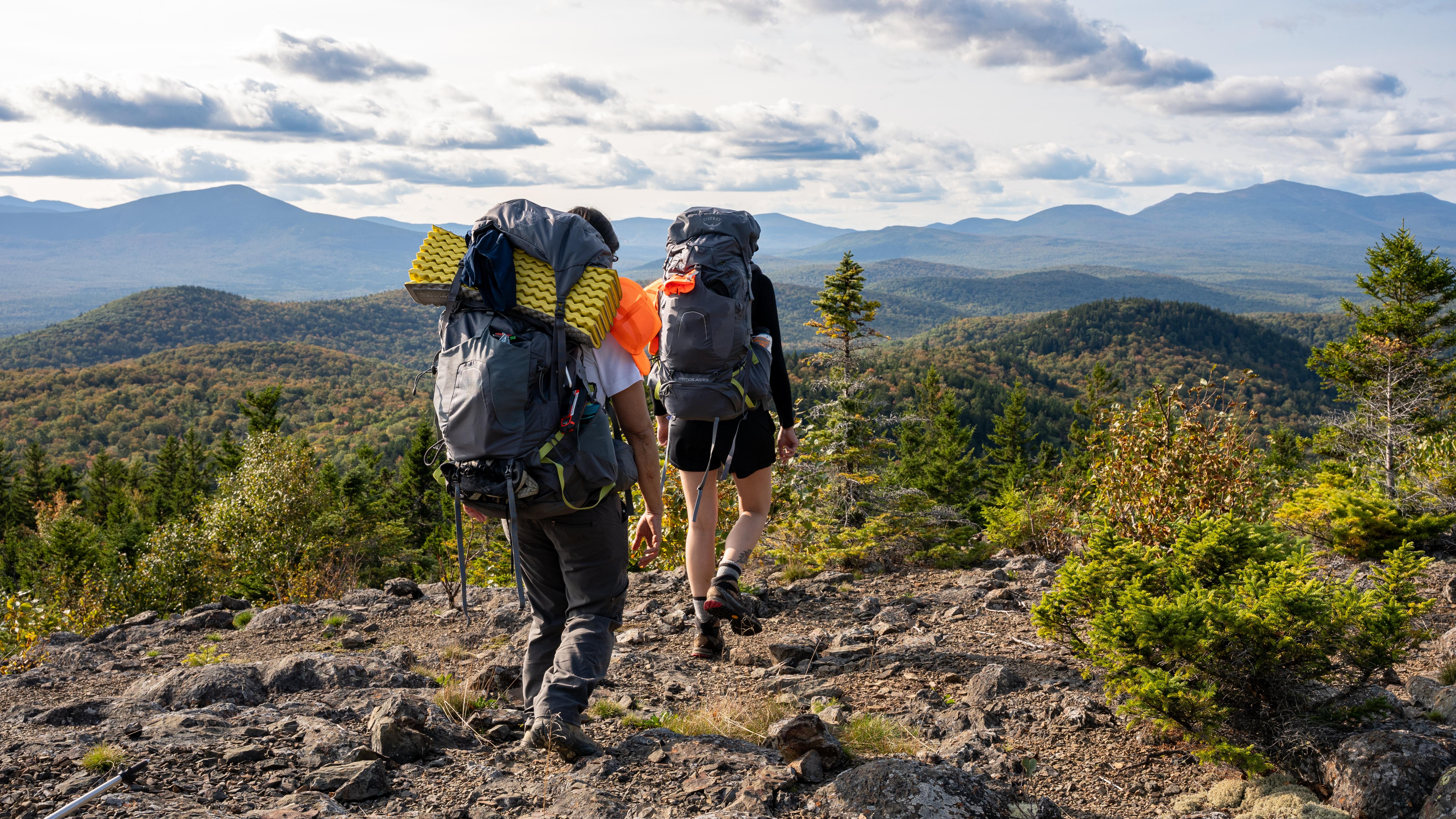 Alt text: Hikers walk across rocky ground. They have backpacks and the background shows mountains.
