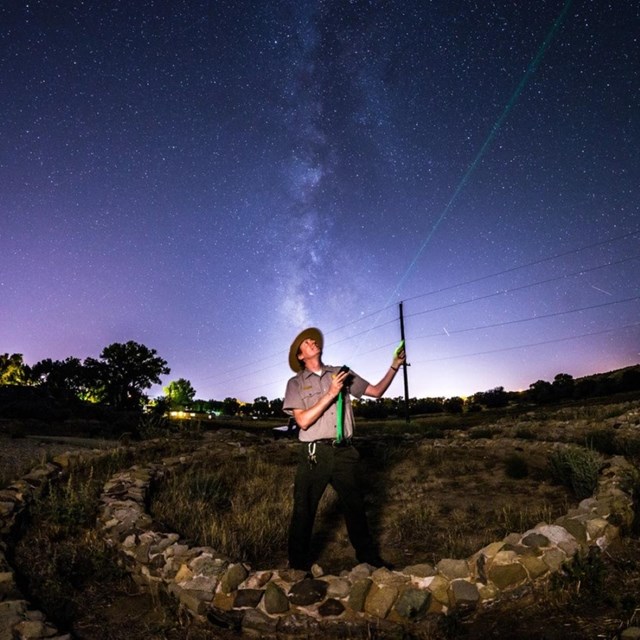 A NPS ranger in uniform holding a measuring instrument up into the night sky.