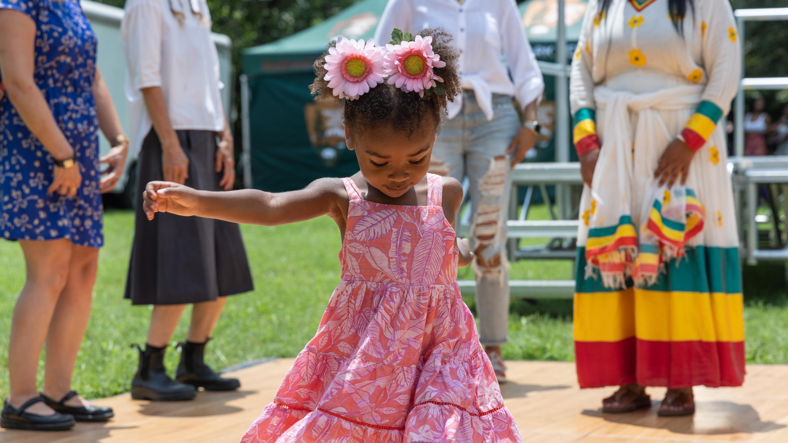 A young girl in a pink dress and floral headband dances as Bethlehem Bekele Belatchew teaches.