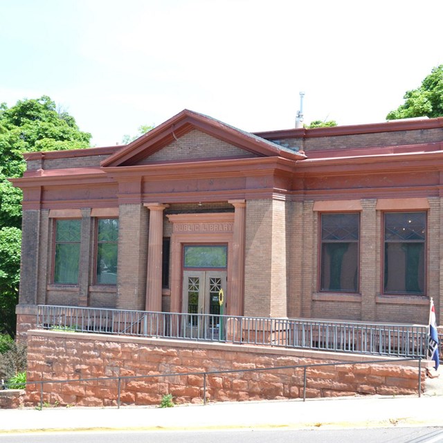 A red brick and stone building sits on a hillside with a street downhill to the left in front.