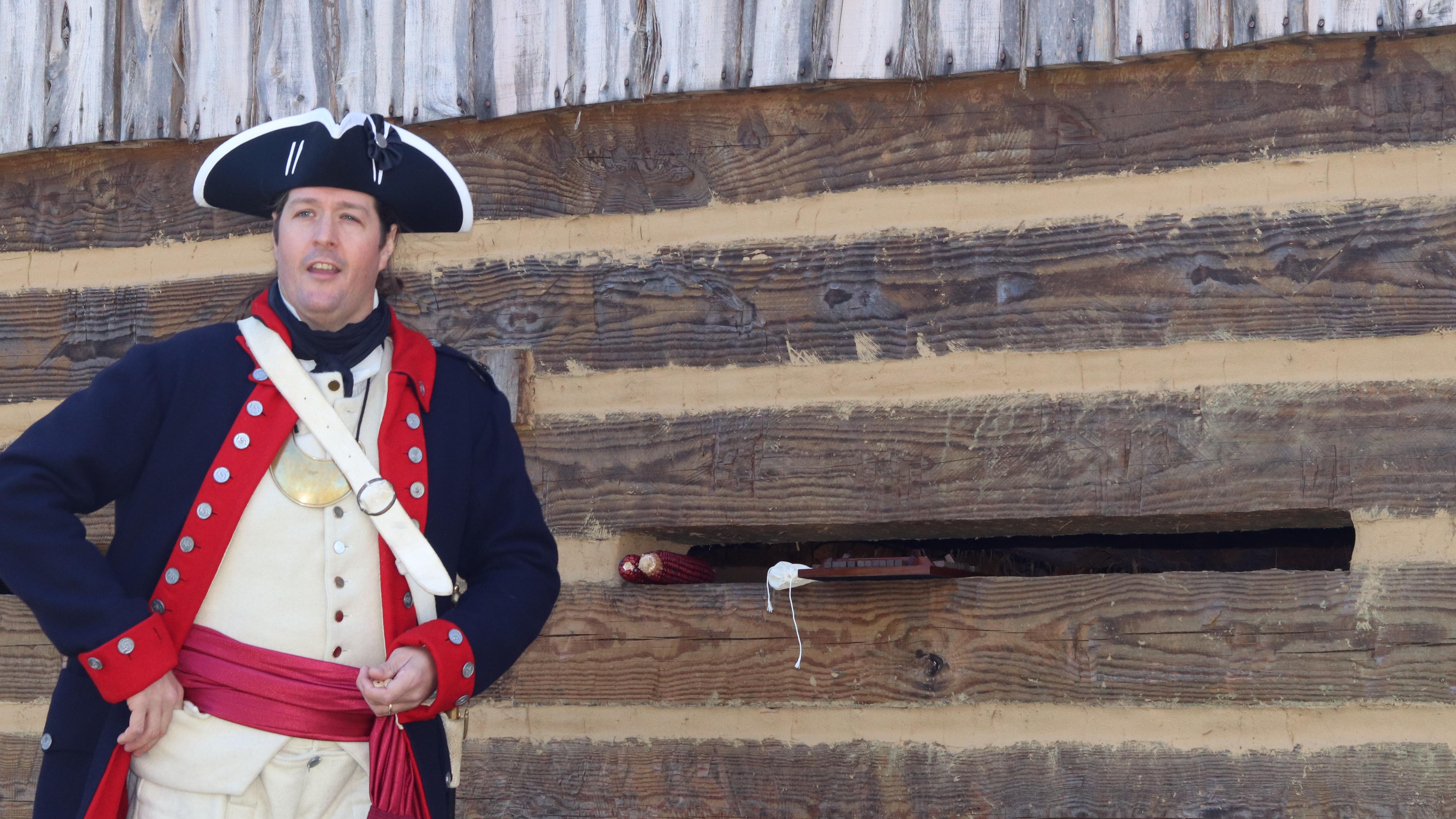 A man in 1700s dress is in front of a wooden building