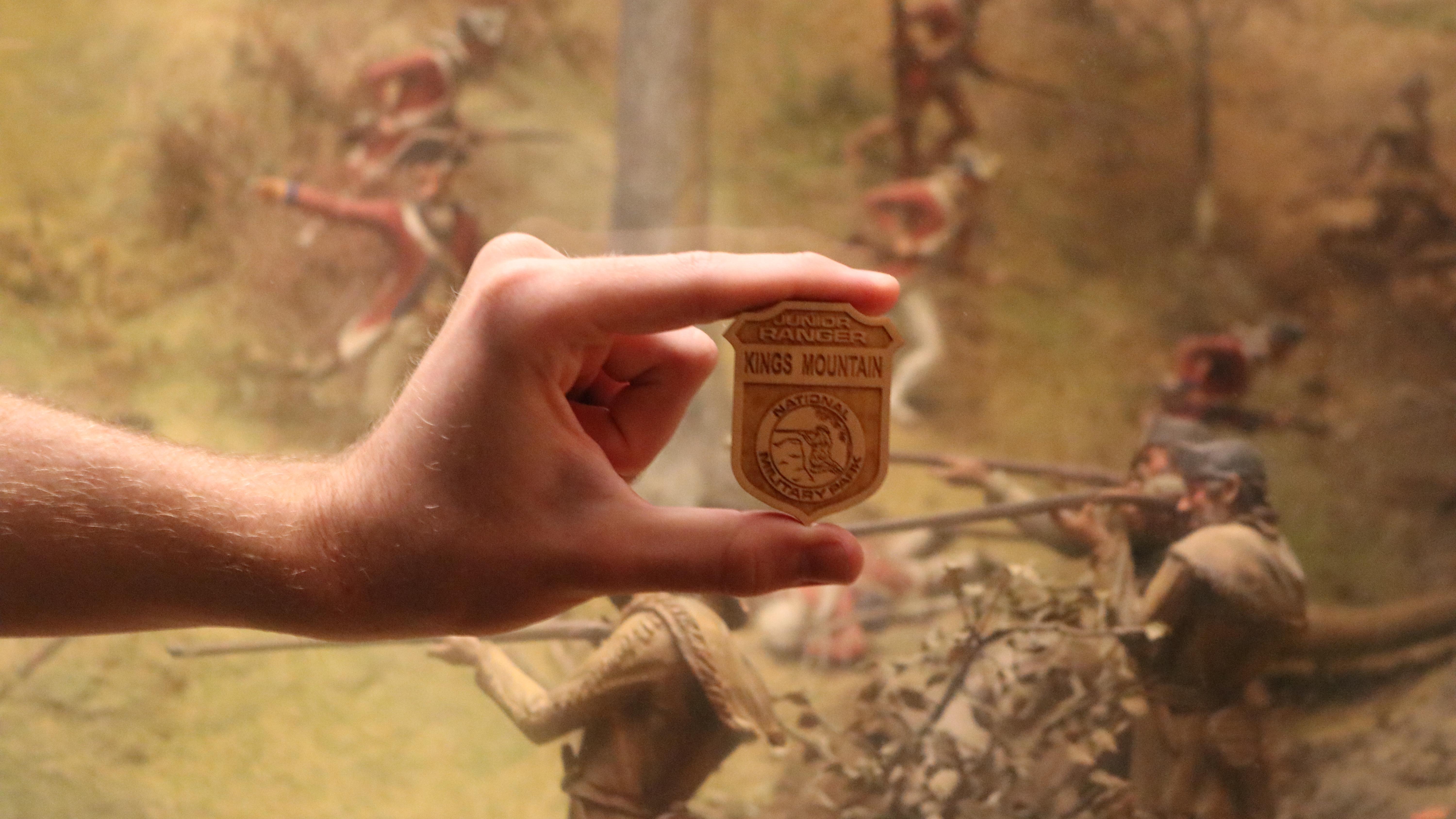 A hand holding a wooden badge that has a soldier firing a musket on it.