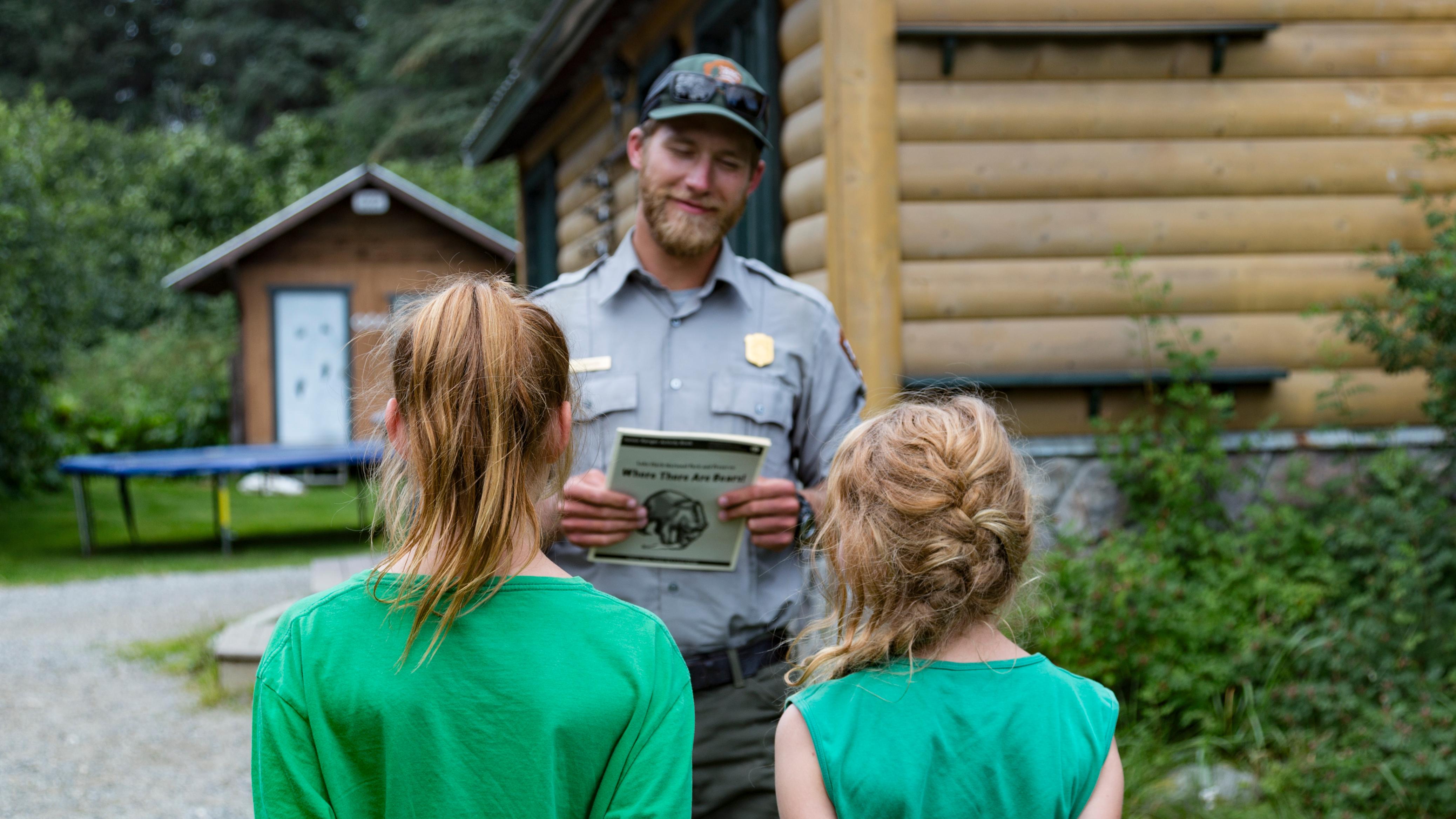 A ranger stands in front of two children in green shirts, holding a Junior Ranger Booklet.