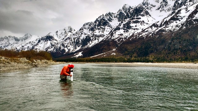 A person stands in a river surveying something, surrounded by mixed-snow mountains. 