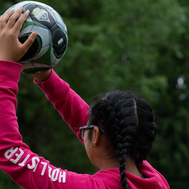 Girl with black braids wears a neon pink hoodie and holds a soccer ball above her head as if she is 
