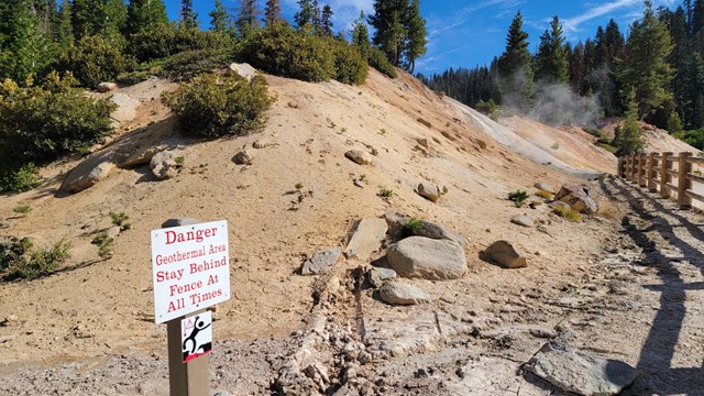 sign reads "Danger, Geothermal Area, Stay behind fence at all times" next to a steaming vent