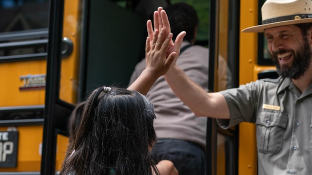 Girl high-fives with a park ranger before boarding school bus.