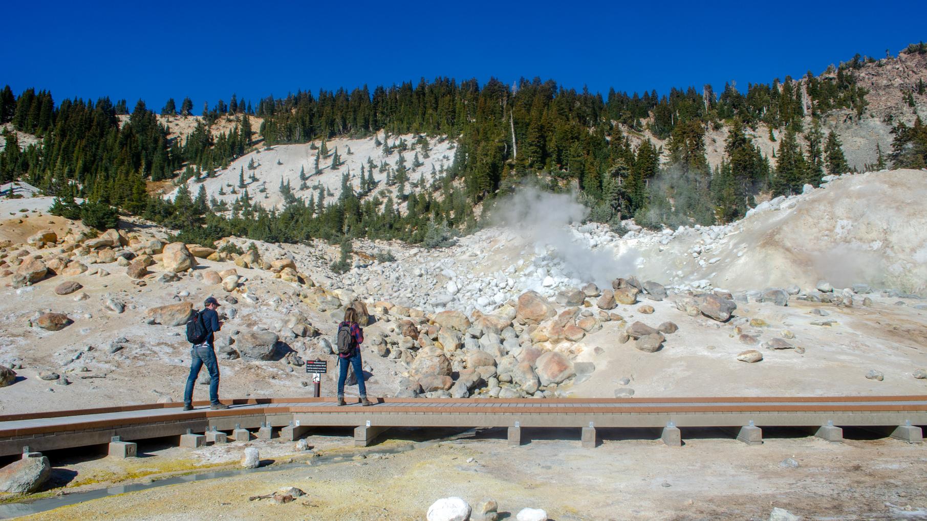 A man and a woman walk along a boardwalk in a hydrothermal area. 