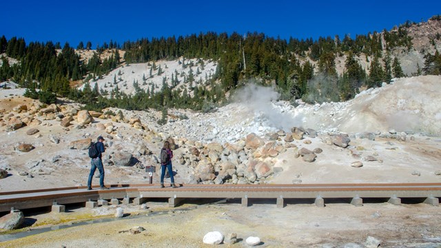 A man and a woman walk along a boardwalk in a hydrothermal area. 