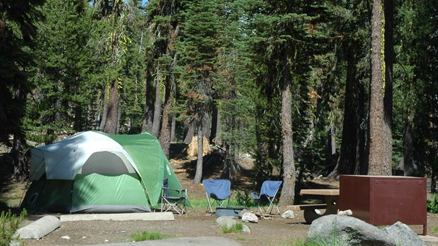 A campsite with a green tent, two chairs in front of a fire pit, a picnic table, and metal box