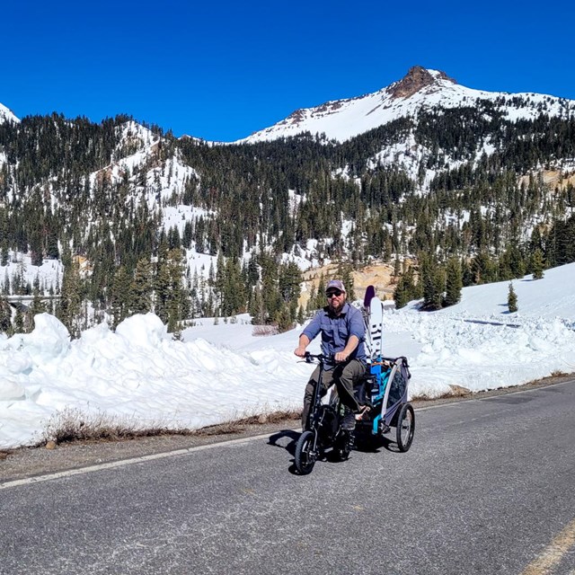 A man rides a small bicycle pulling a trailer on a road lined by snow.