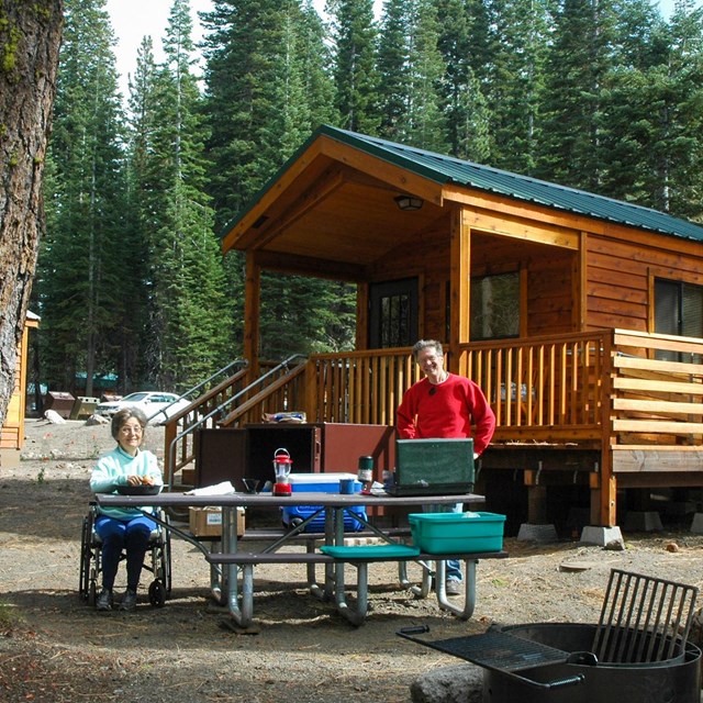 A man stands and a woman sits in a wheelchair at either end of a picnic table in front of a wooden c