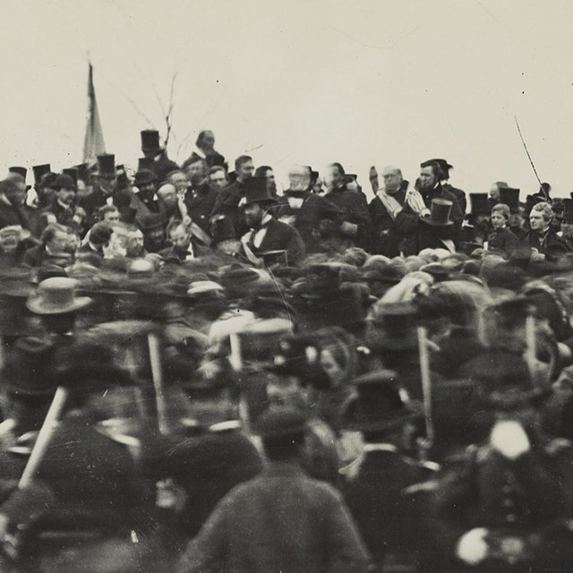Crowd of people around Abraham Lincoln who stands at podium, giving a speech