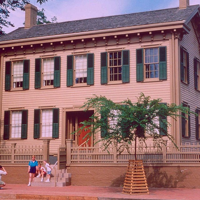 Photo of Lincoln Home, a 2 tan story wooden house with darker trim, green shutters, and 2 chimneys
