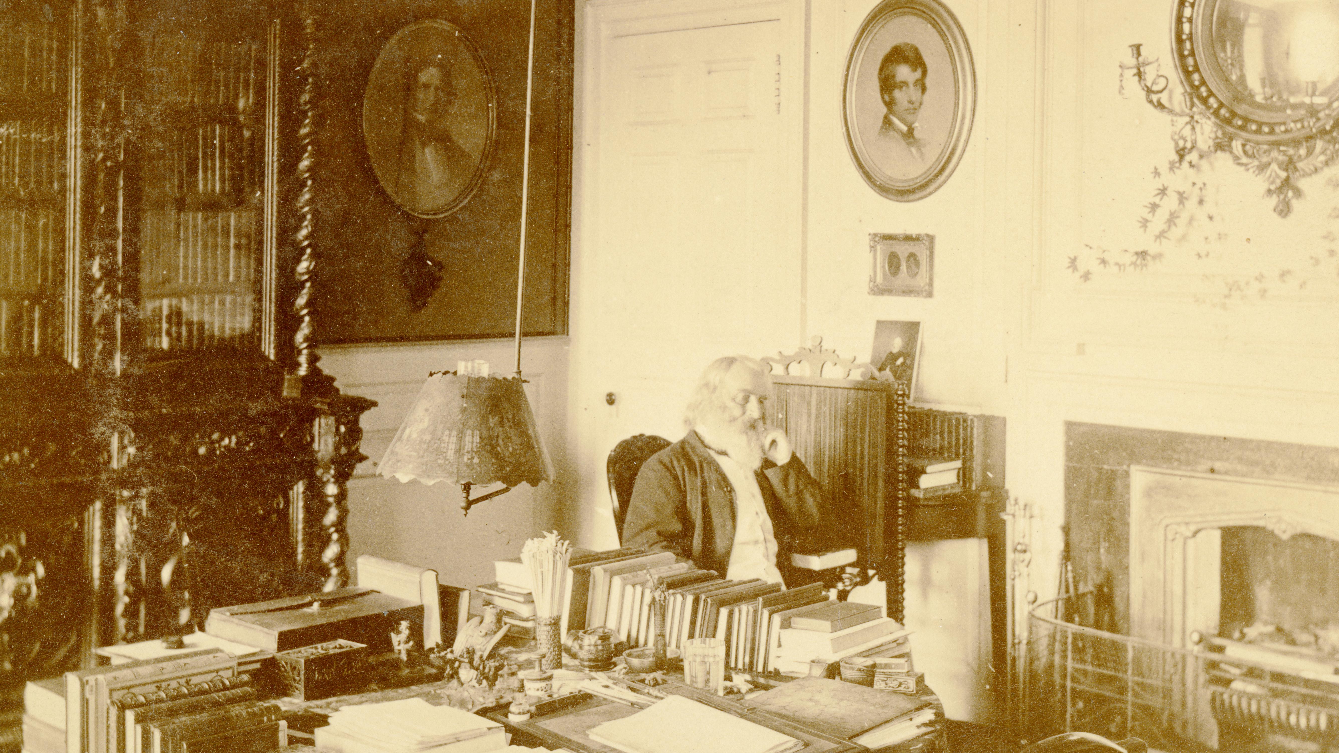 black and white photograph of man seated at table covered in books