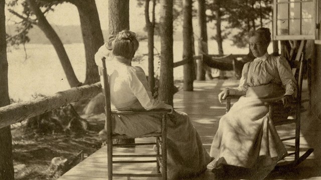 Two women seated on long rustic porch, looking over lake. One woman faces towards the camera in a ro
