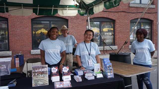 Four people smile for the camera all wearing volunteer shirts at a table with brochures and info.