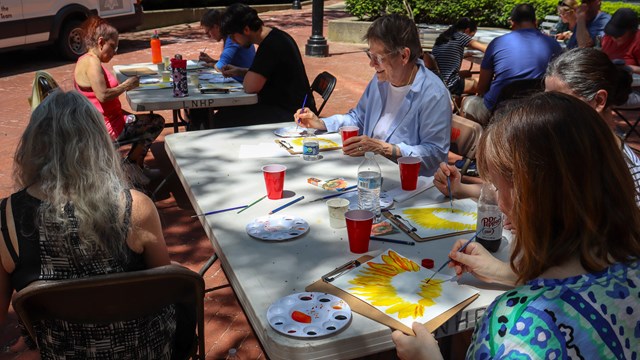 A group of people sit around tables painting sunflowers and smiling.