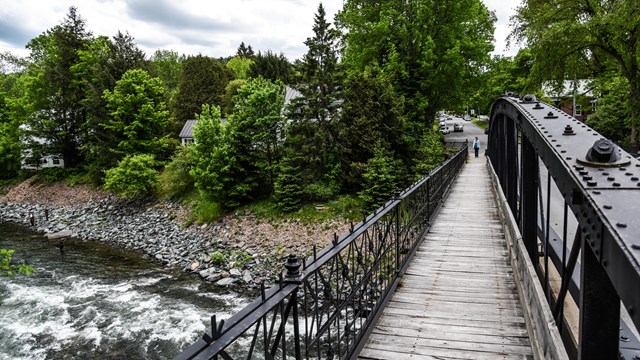 An iron bridge with a wooden footpath crosses over a river with lush green trees in the background