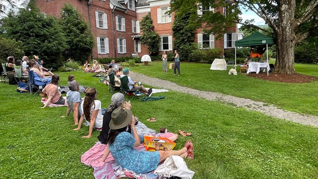 group of people in lawn chairs watch a performance in front of a large red brick mansion