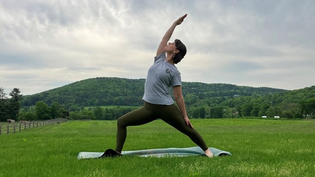 Woman in NPS shirt does a yoga pose with a green mountain in the background