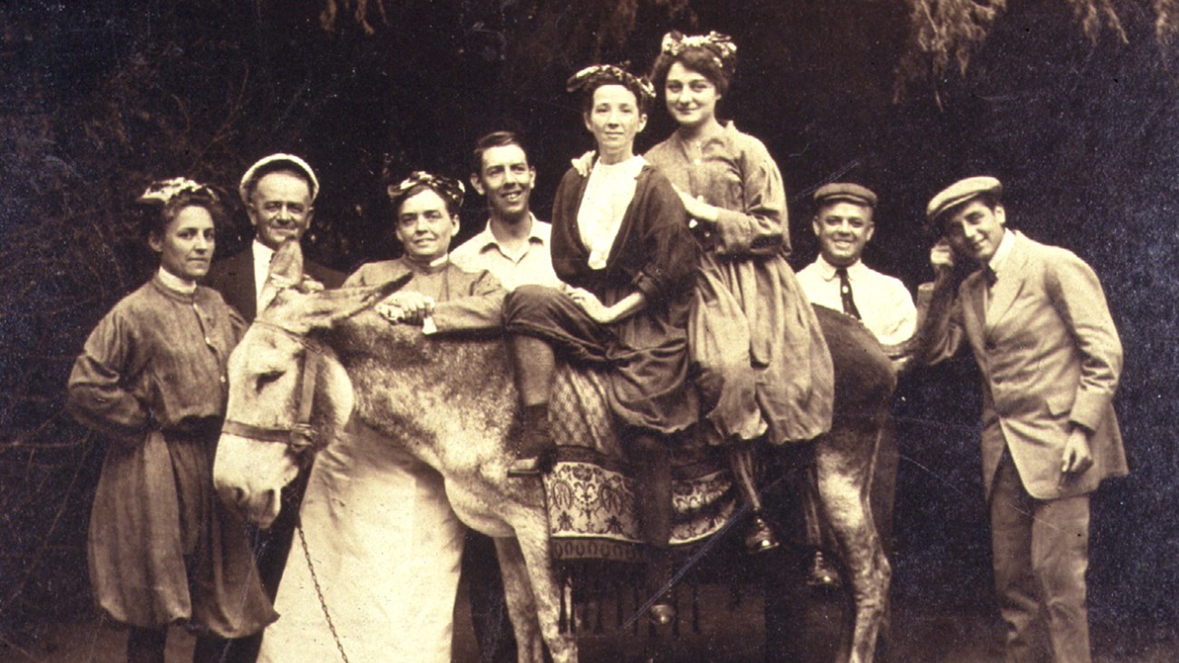 A group of people wearing clothing from the 1900's who are standing around and sitting on a donkey.