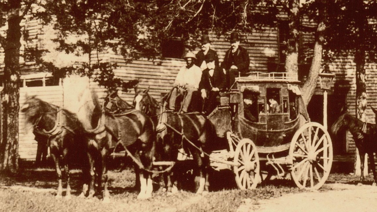 Black and white photograph of a large four horse drawn stagecoach in front of a long white building.