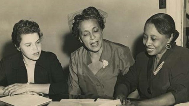 Three women sit at a desk talking about news.