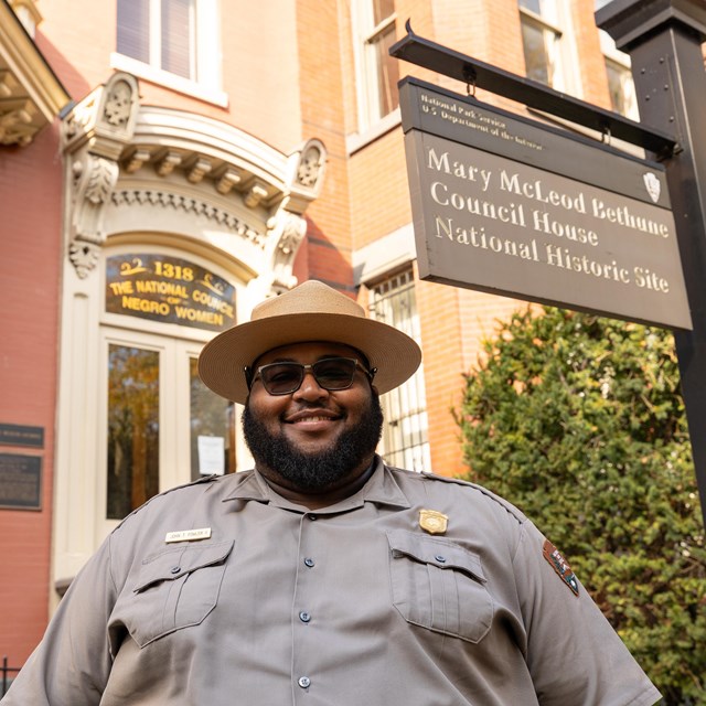A park ranger smiles at the camera in front of a historic home