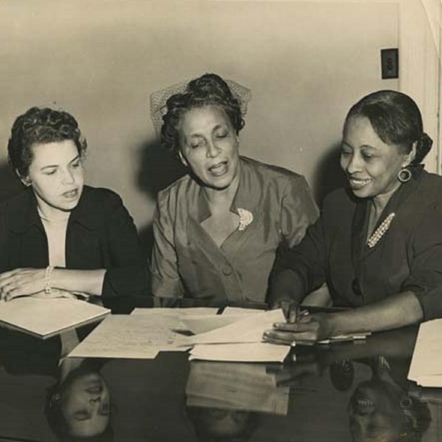 Three women sit at a desk talking about news.