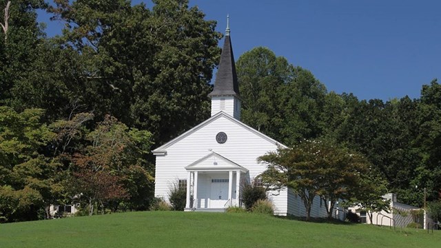 A small white chapel with a tall steeple on a grassy hill with trees behind it and an historical mar