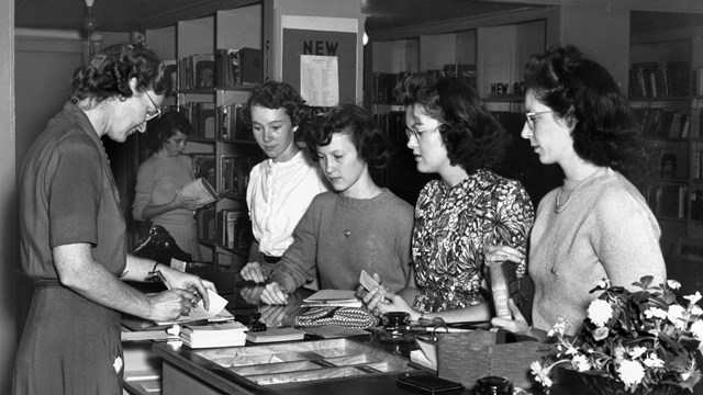 4 young women stand at a counter. The woman behind the counter writes on a card.