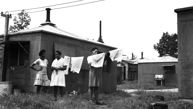 A black and white photo of women near laundry and small dwellings.
