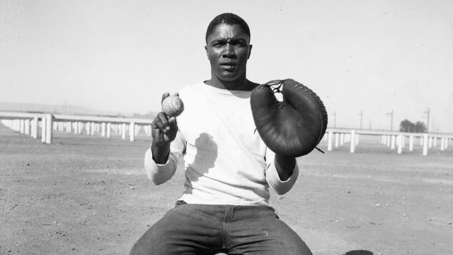 A black and white photo of a black man in a white t-shirt and dark jeans holding a baseball glove an