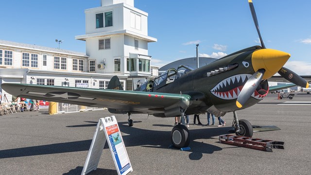  An image of a World War II-era fighter plane on a runway with buildings behind it.